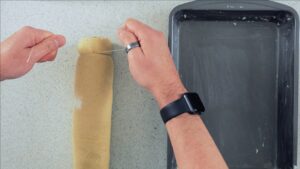 Roll of cinnamon roll dough being cut by unwaxed, unflavored dental floss near a prepared pan.