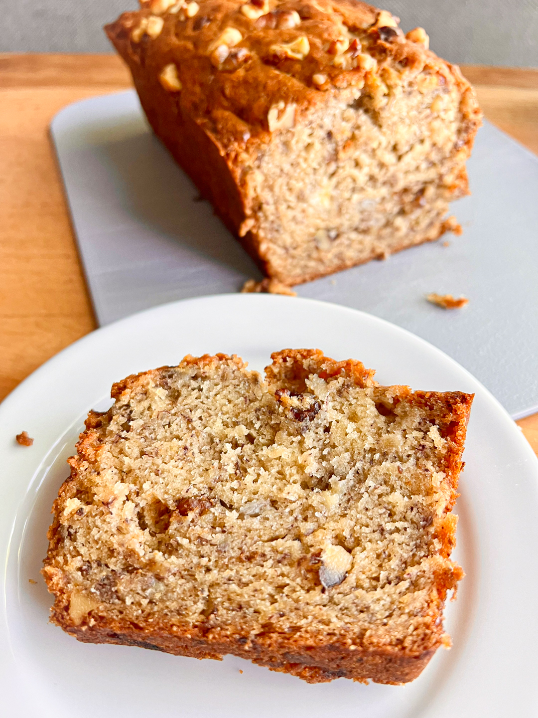 This is an image of a banana bread slice on a white plate with a loaf of banana bread in the background.