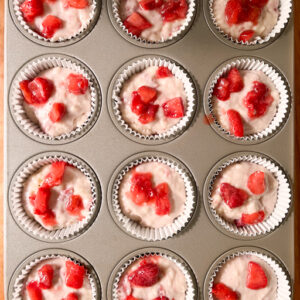 An image of batter-filled muffin cups with strawberries sprinkled on top.