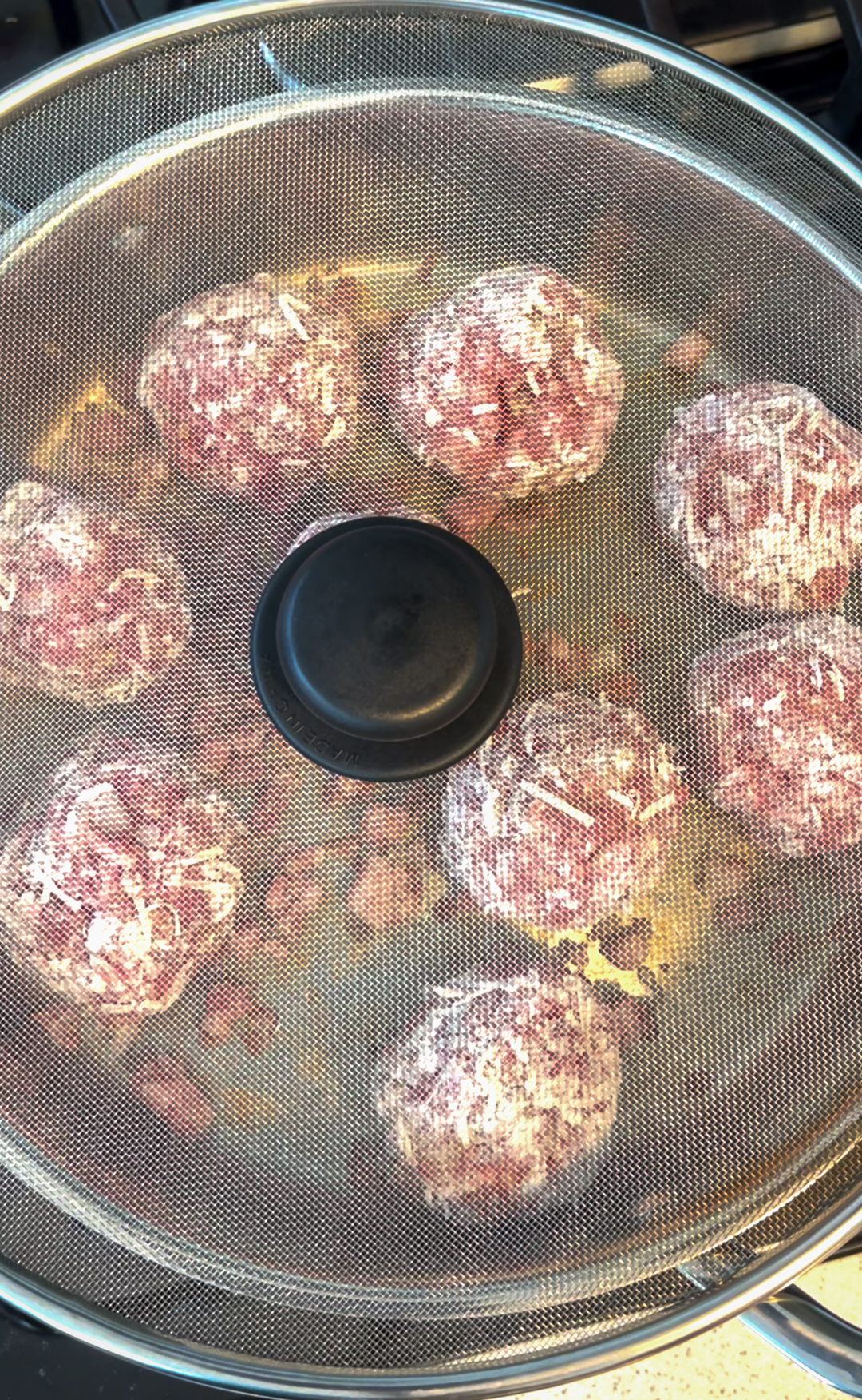 This is an image of meatballs being cooked in a stainless steel pan with a splatter screen placed over it.