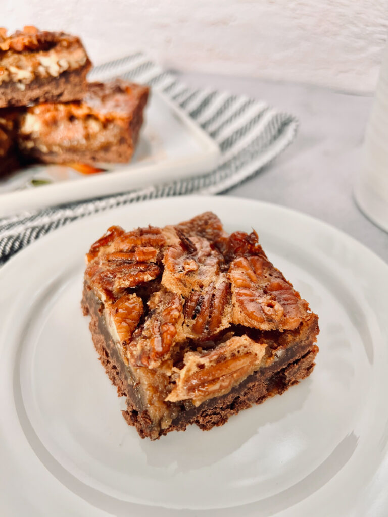 Chocolate pecan pie bar on a white plate, with additional pecan pie bars blurred in the background.