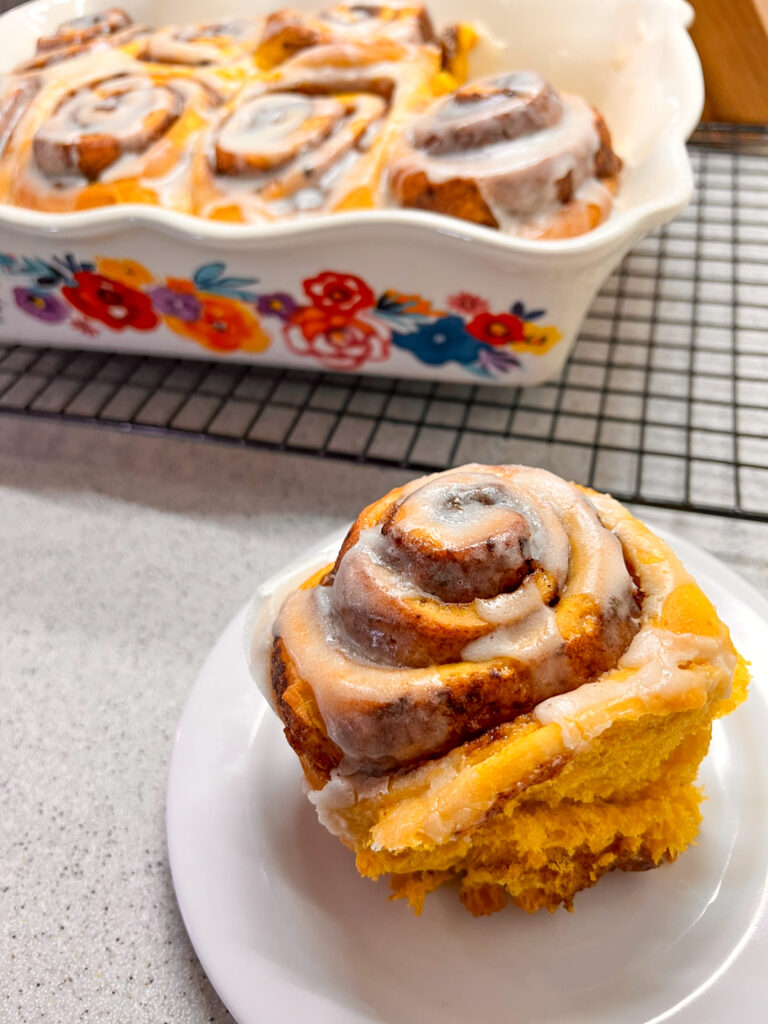 A pumpkin spice latte cinnamon roll on a white plate with additional cinnamon rolls blurred in the background.